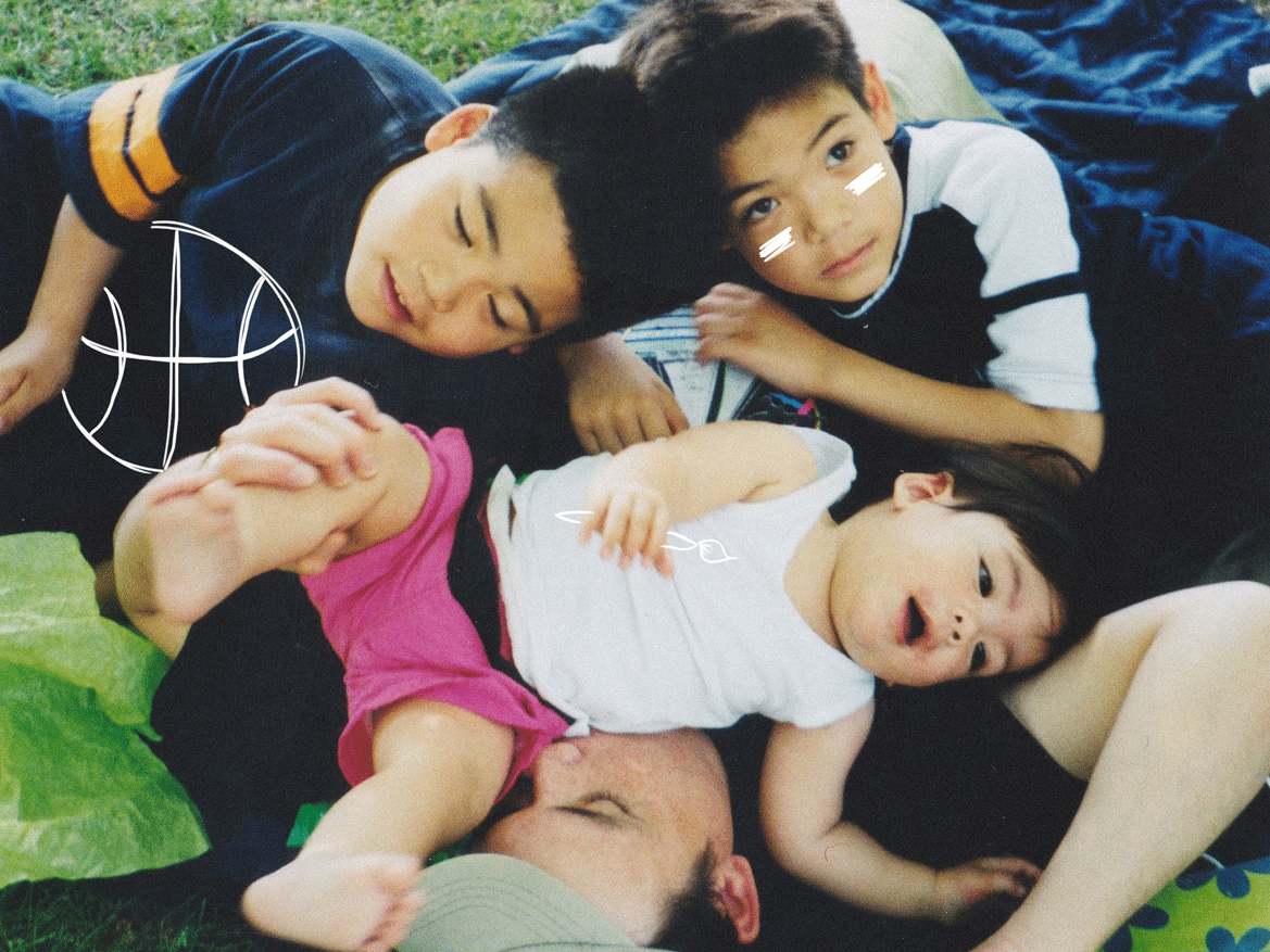 A dad laying in the grass with his three sons piled on top of him at Assiniboine Park in the year 2001. A doodle animation puts a paintbrush, basketball, and football makeup in the photo.